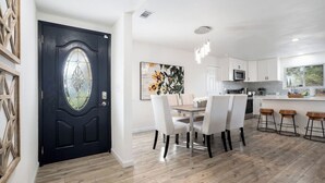 Eine moderne Küche mit weißen Schränken, eine Essecke mit einem Holztisch und weißen Stühlen sowie eine schwarze Tür mit Fenster.
