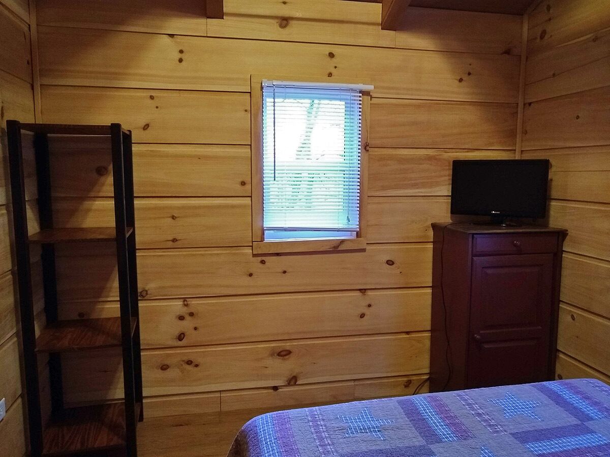 A wooden cabin room with a bed, a small window, a TV on a cabinet, and a ladder.