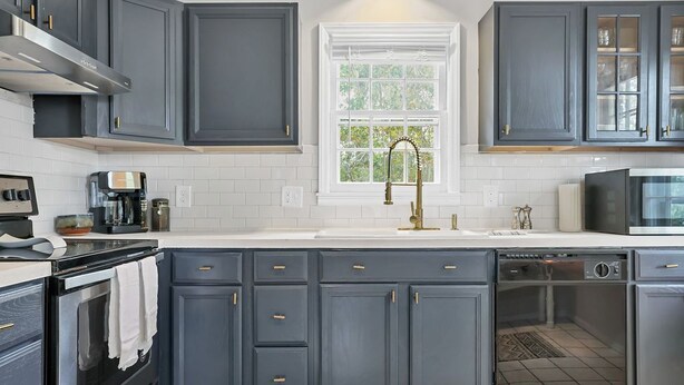 A modern kitchen with dark grey cabinets, a white countertop, a stainless steel range hood, a built-in dishwasher, and a window with a view of greenery. - HEART OF DOWNTOWN (Riverwalk, River View, Walk to Shopping, Dining, and Spirits) (Wilmington)