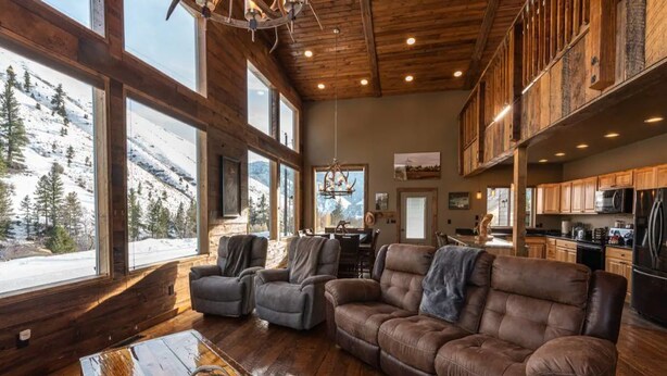 Ein geräumiges Wohnzimmer mit großen Fenstern mit Blick auf einen verschneiten Berg, Holzböden, Ledersofas und einer rustikalen Decke.