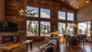 Ein gemütliches Wohnzimmer mit Kamin, großen Fenstern mit Blick auf einen verschneiten Berg und einem Essbereich mit Tisch und Stühlen, sowie ein Ledersofa.