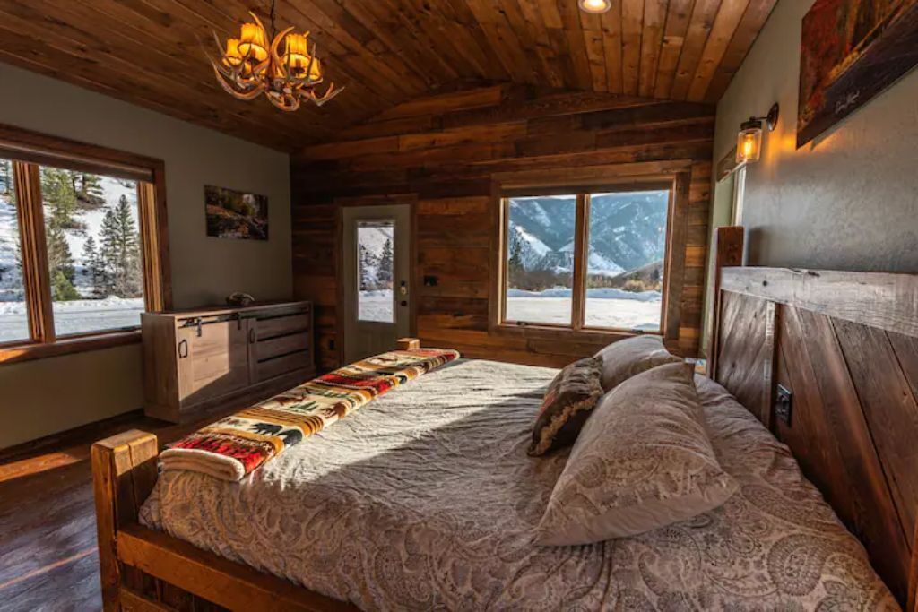 A cozy wooden cabin bedroom with a large bed, a dresser, and a view of snowy mountains through the windows.