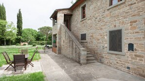 Un bâtiment en pierre avec un escalier menant à un balcon, des sièges à l’extérieur et une zone herbeuse.