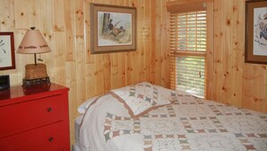 Una stanza con pannelli di legno, un letto, una cassettiera rossa, una lampada e due quadri appesi alle pareti.