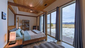 Een slaapkamer met een groot raam, houten balken in het plafond, een bed met een lichtkleurig hoofdeinde en een nachtkastje met een lamp.