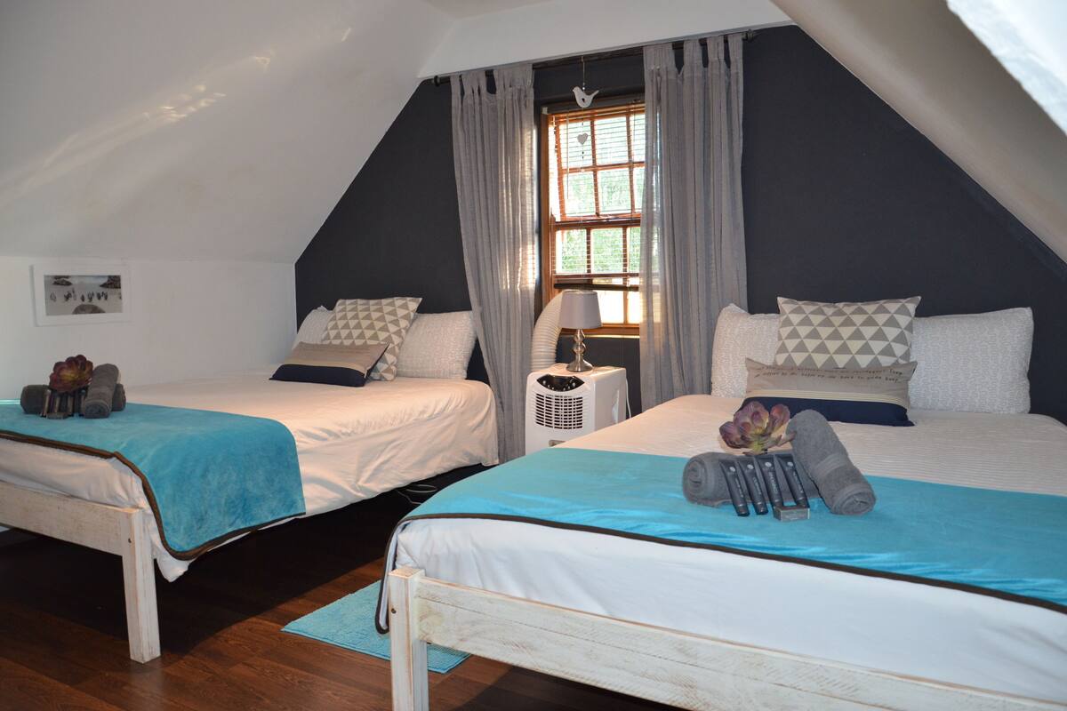 A bedroom with two beds, a window with curtains, a bedside table with a lamp, and a small heater.
