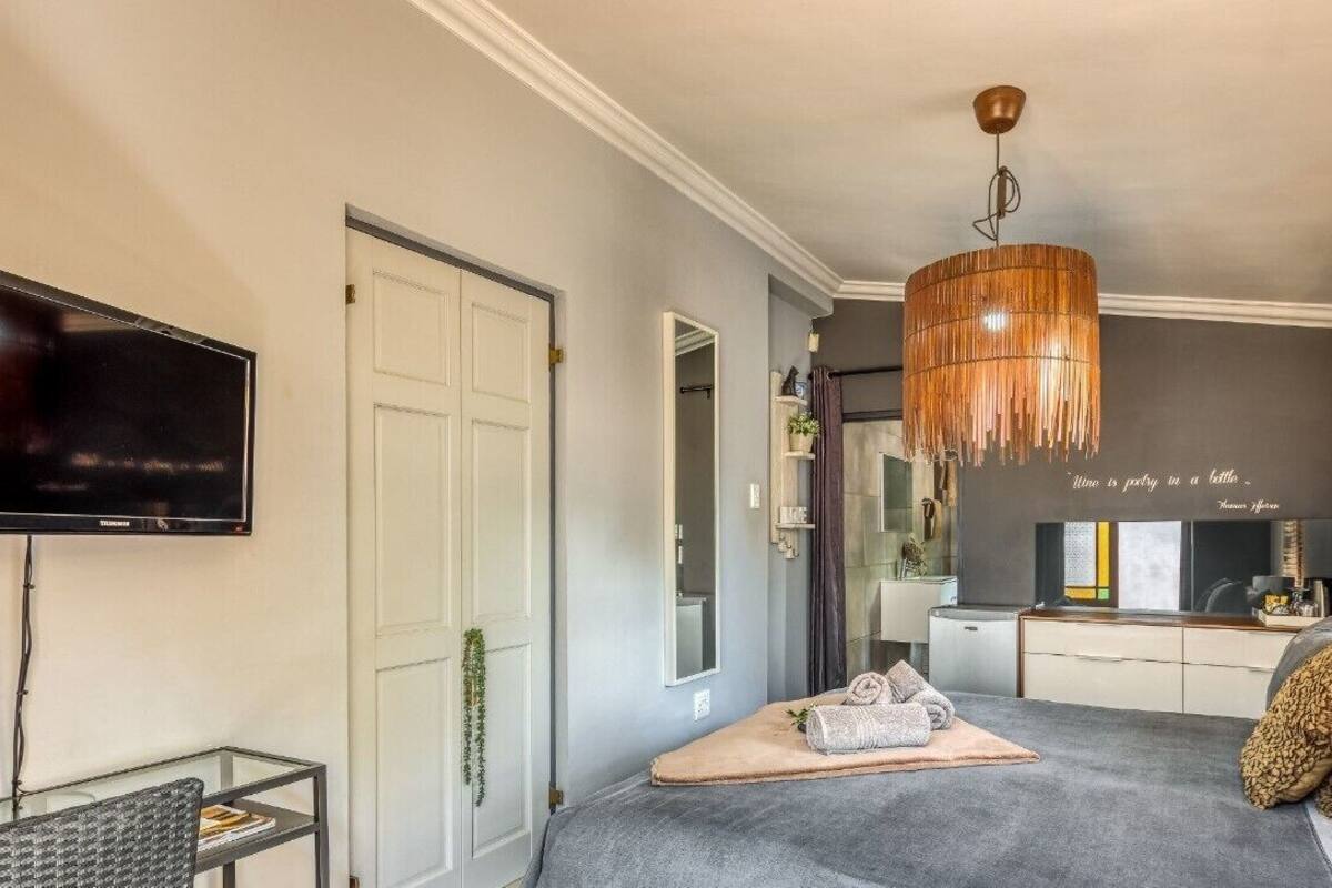 A modern bedroom with a flat-screen TV, a glass coffee table, a bed with a beige throw, a hanging wooden light fixture, and a bathroom visible in the background.