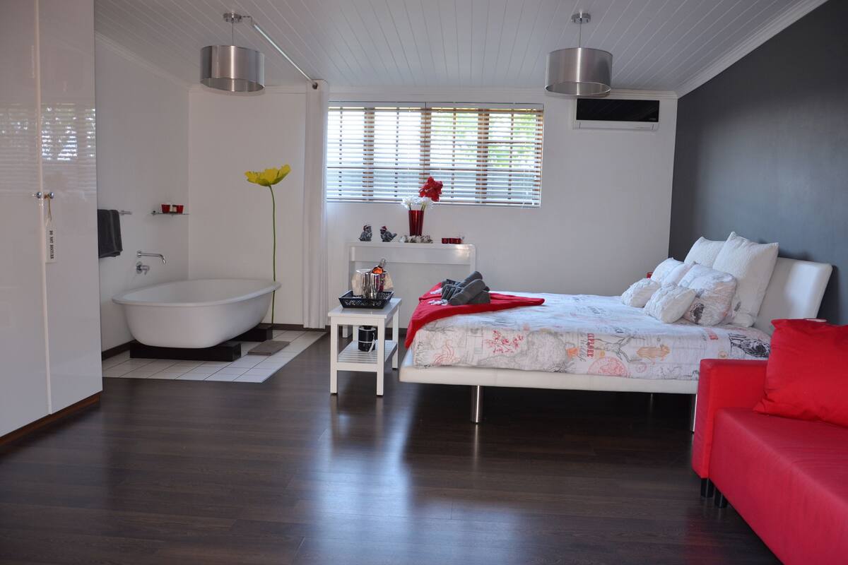 A modern bedroom with a large bed, a red sofa, a bathtub, and a window with blinds.