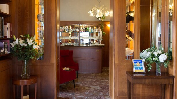 Un espace bar avec un comptoir en bois, des étagères remplies de bouteilles et de verres, et un fauteuil rouge. De chaque côté de l’entrée, on trouve des vases ornés de fleurs.