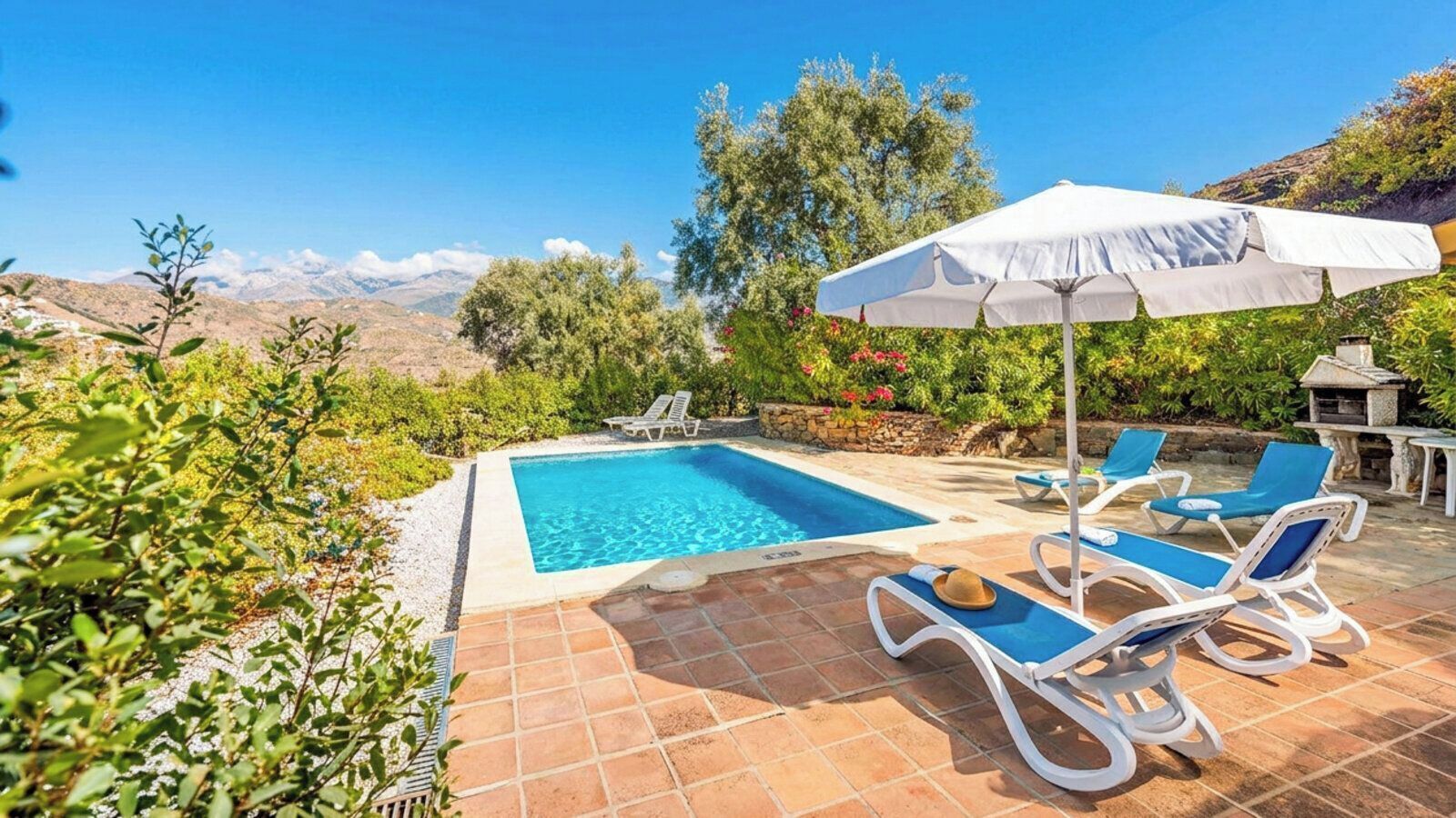 A pool area with a clear view of mountains in the background, surrounded by greenery and equipped with lounge chairs and a sun umbrella.