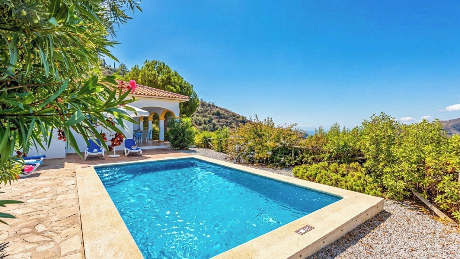A pool area with a clear blue pool, a small building with a tiled roof, and a view of the surrounding landscape.