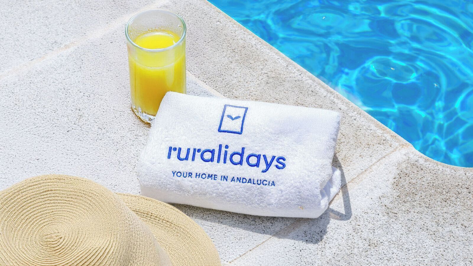 Una toalla con la marca 'ruralidays', un vaso de zumo de naranja y un sombrero de paja junto a una piscina.