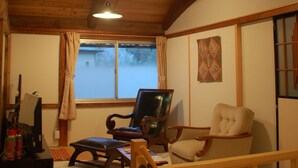 Habitación con techo de madera, ventilador de techo, ventana con cortinas, una silla, una mesita y televisor.
