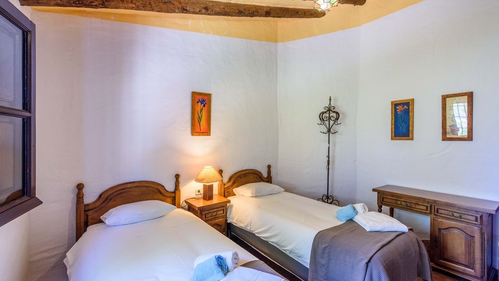 Una habitación con dos camas de madera, una mesita de noche con lámpara, un escritorio de madera y un perchero en la pared.