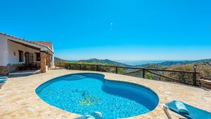 Ein Pool mit einer Steindeck, einem Holzzaun und Blick auf das Meer und die Hügel.