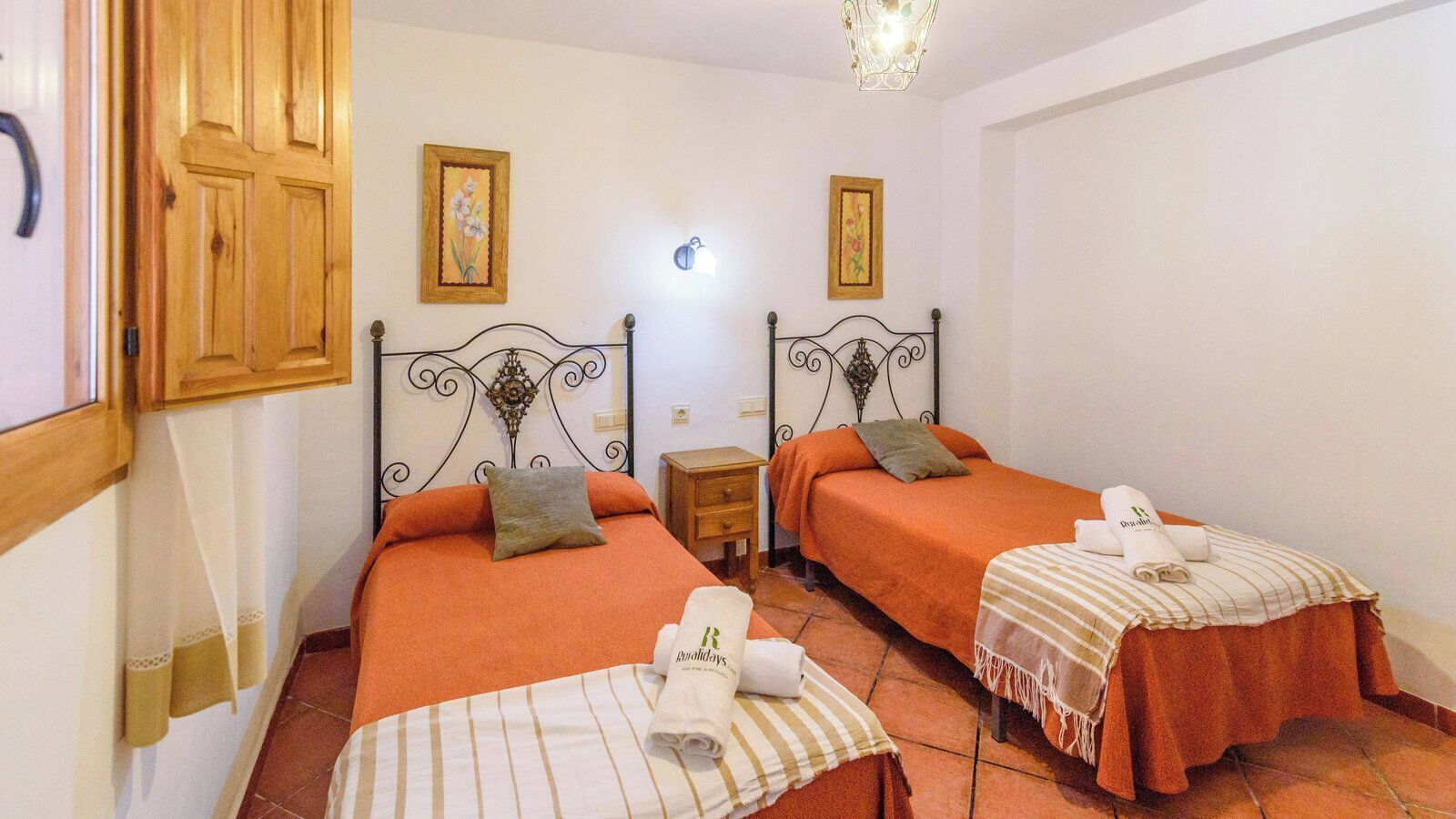 A room with two beds, each with an orange bedspread and a matching pillow. There are two nightstands, one between each bed. The room has a tiled floor and a wooden door.