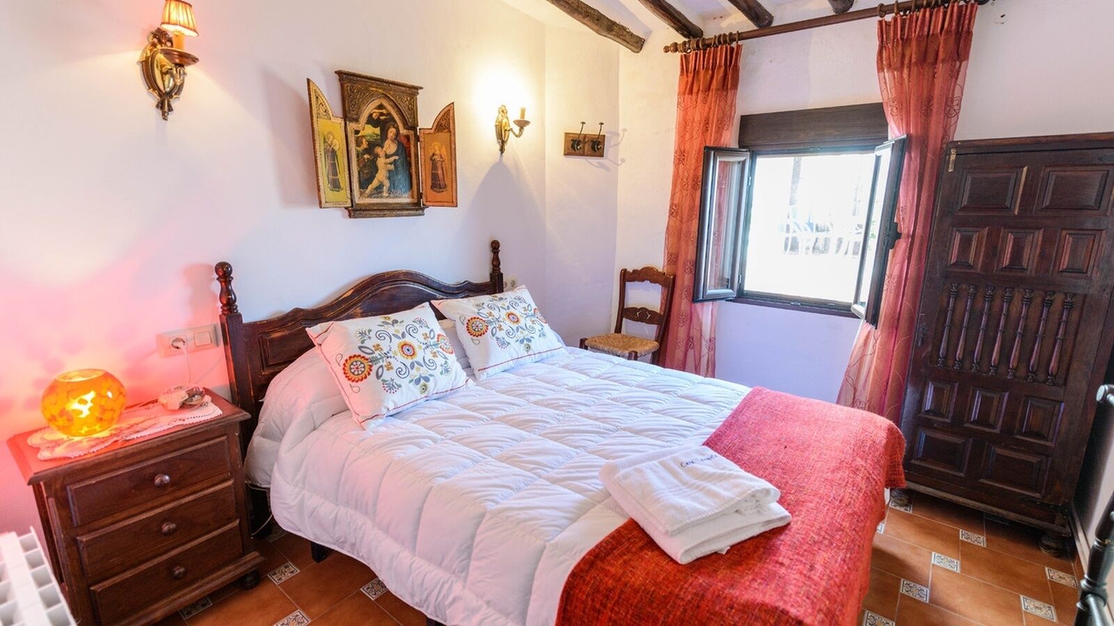Un lit bien fait, avec une couette blanche et des coussins décoratifs, une table de chevet en bois avec une lampe, un tableau religieux accroché au mur et une grande porte en bois.