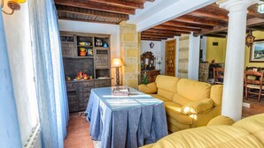 Un salón acogedor con techo de madera, un sofá amarillo, una mesa con lámpara y una estantería con objetos coloridos.