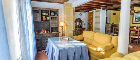 Un salón acogedor con techo de madera, un sofá amarillo, una mesa con lámpara y una estantería con objetos coloridos.