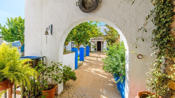 Un sendero arqueado que conduce a un edificio de fachada blanca y detalles azules, rodeado de macetas con plantas y una campana en la pared.