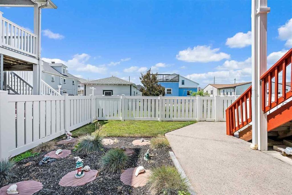 Un jardin à l’arrière d’une maison, avec une clôture blanche, un chemin en béton et un petit espace aménagé en jardin, agrémenté de pierres plates servant de pas.