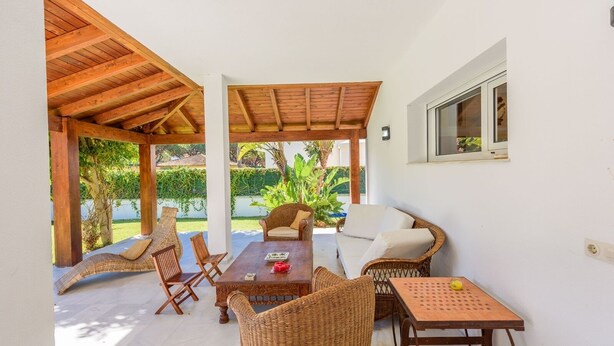 A covered outdoor seating area with wicker furniture, a wooden table, and a view of a garden. - Villa con Encanto Conil de la Frontera by Ruralidays (Cádiz)