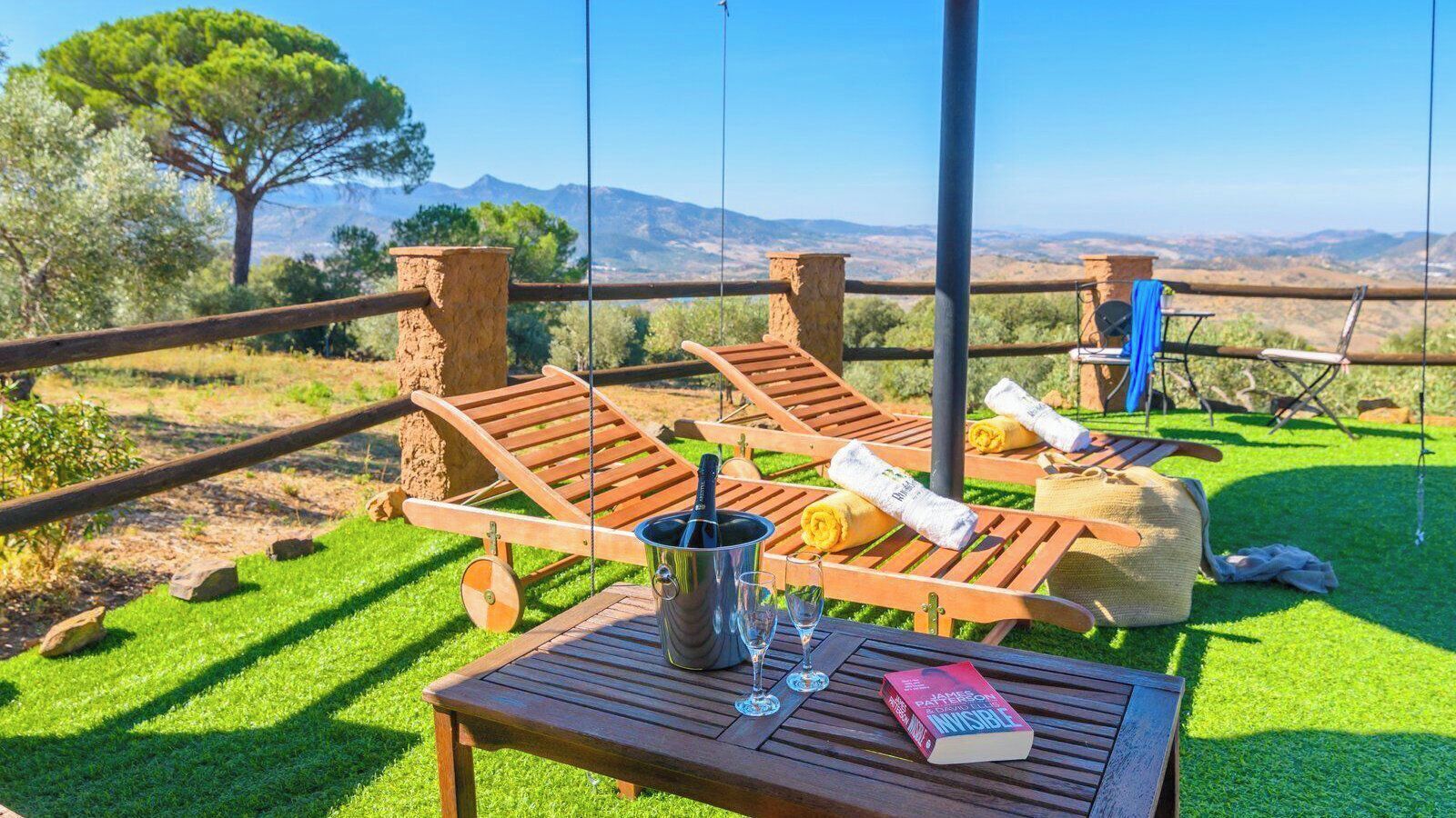 Eine Terrasse mit Holzliegen, einem Tisch mit Flasche, Gläsern und einem Buch, mit Blick auf Berge.