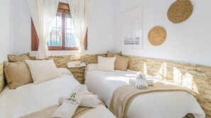 A hotel room with two beds, a stone wall, a window with curtains, a wall-mounted lamp, and a framed picture. - La Dehesa de Torrox by Ruralidays (Málaga)