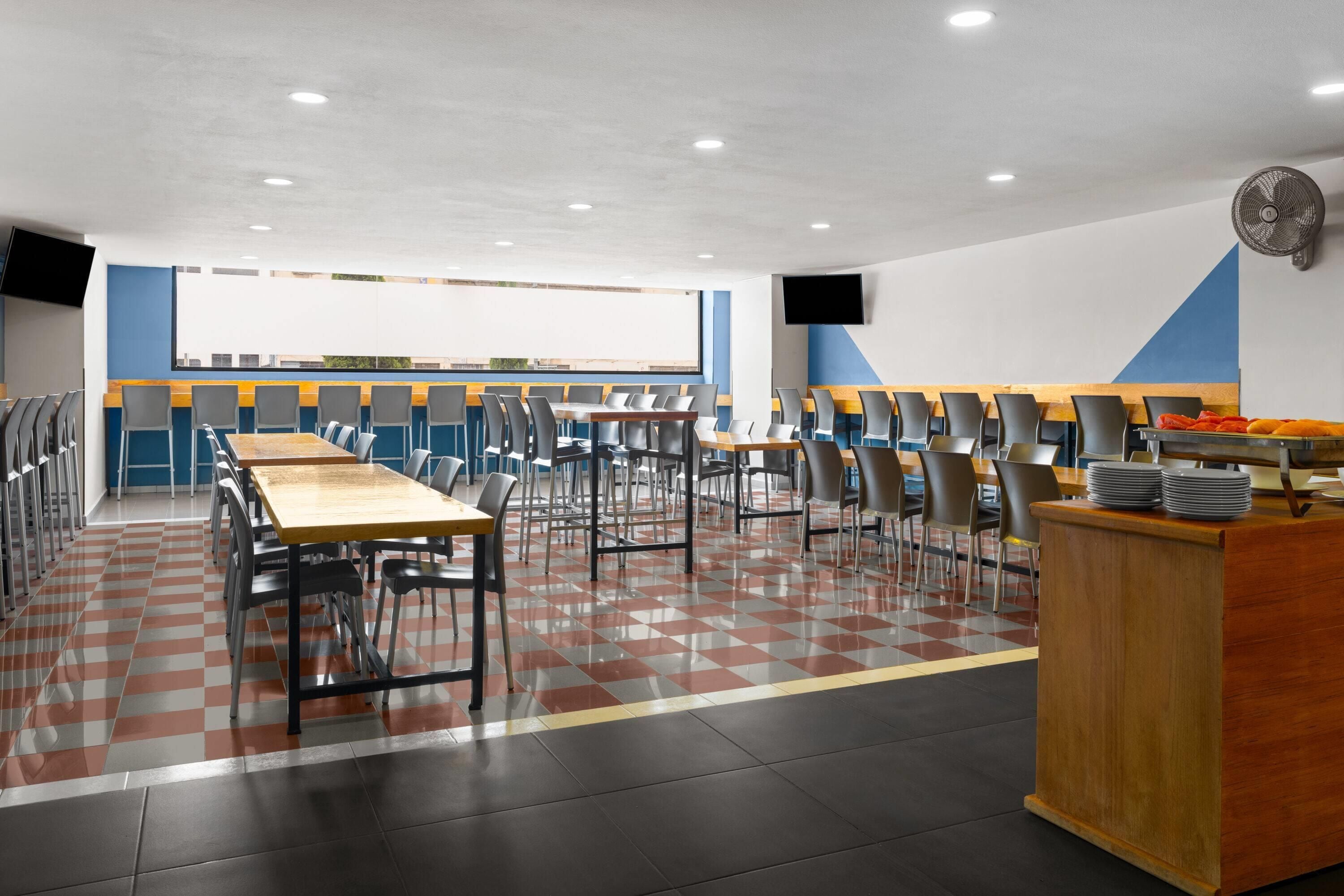 Una cafetería con varias mesas y sillas, un mostrador de madera con platos, y un amplio ventanal al fondo.