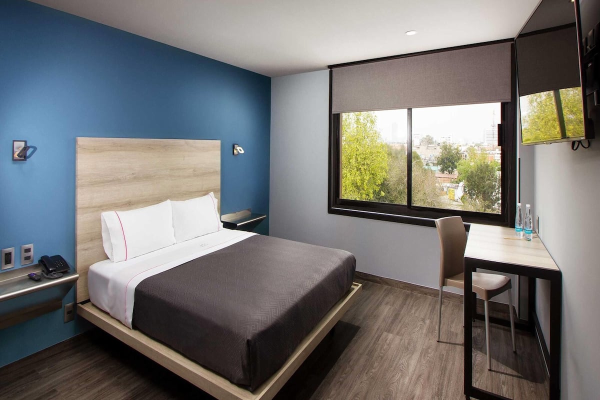 Een moderne hotelkamer met een bed, bureau en stoel, een raam met uitzicht op de stad, en een blauwe accentmuur.