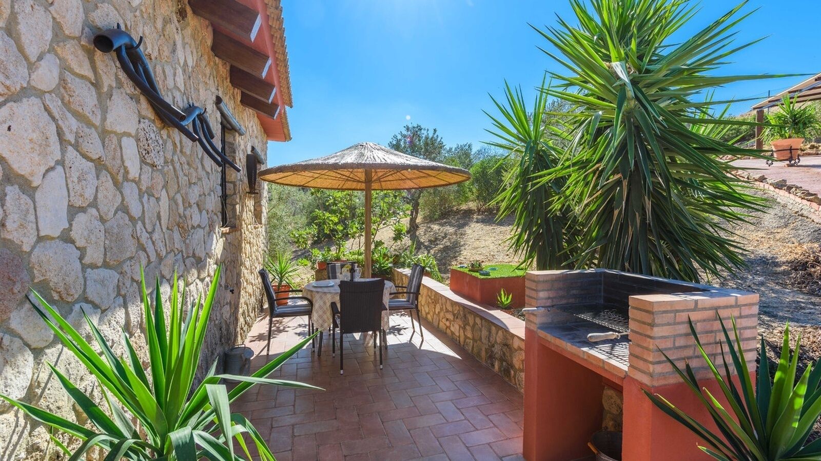 Une terrasse avec des murs en pierre, une table avec des chaises, un barbecue et un grand parasol.