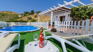 Zona junto a la piscina con una pérgola blanca, una mesita con una botella y vasos, y una silla de descanso.