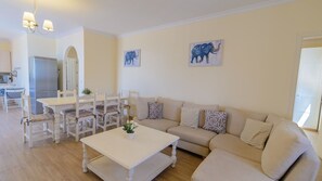 Una sala de estar con un sofá color beige, una mesa de centro blanca y un área de comedor con una mesa y sillas blancas. Hay dos cuadros de elefantes colgados en la pared.