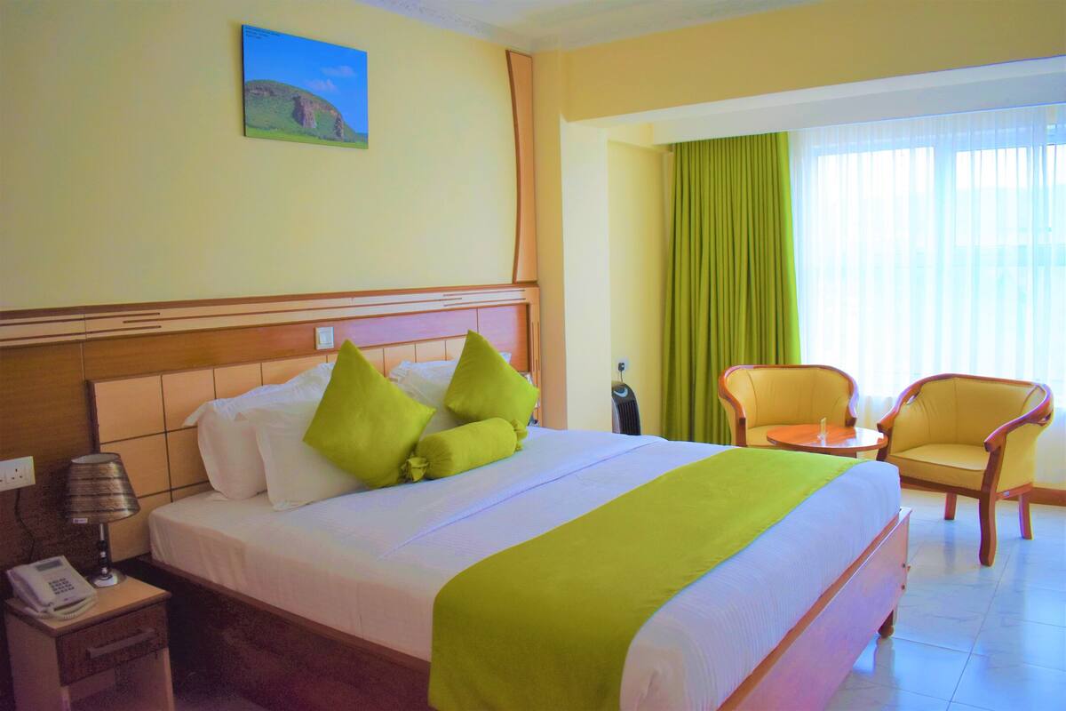 Een hotelkamer met een bed, nachtkastje met telefoon, stoel en raam met gordijnen.