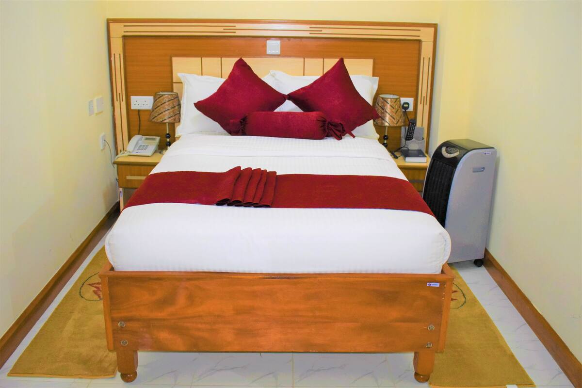 Een hotelkamer met een groot bed, een houten hoofdbord, twee rode kussens, een nachtkastje met een lamp, een telefoon en een bureau met een computer.