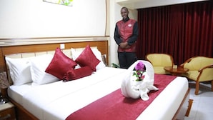 A hotel room with a neatly made bed, a red throw blanket, and a floral arrangement on the bed. There is a wooden headboard, a nightstand with a glass, and a chair in the corner.