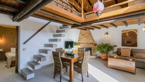Ampla sala de estar com escada de madeira, lareira e mesa de jantar com cadeiras estampadas.