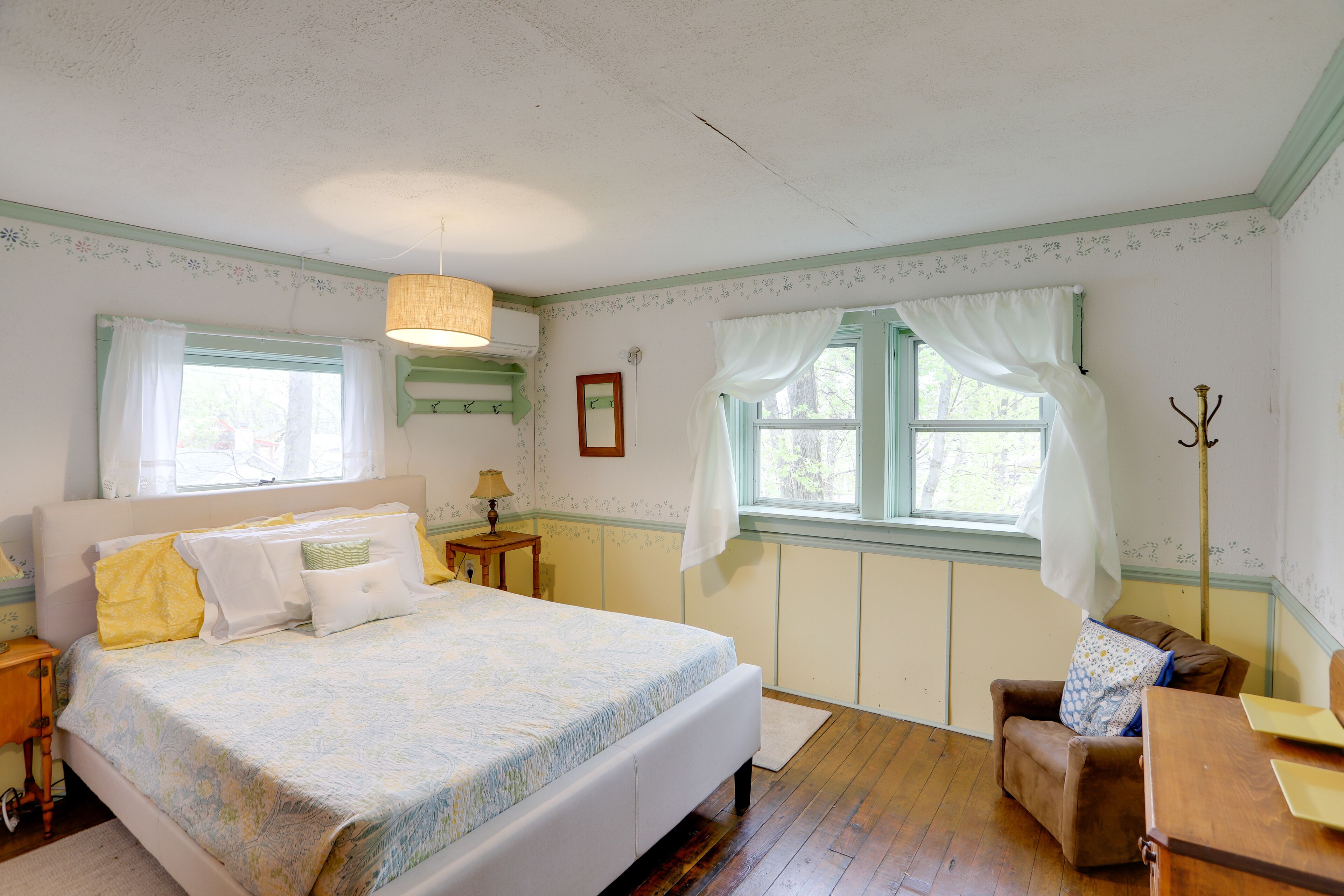 Ein Schlafzimmer mit einem großen Bett, zwei mit Vorhängen versehenen Fenstern, Holzboden und einem Sessel.
