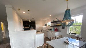 Cozinha moderna com armários brancos, área de jantar com mesa branca e vista para o exterior através de uma janela.