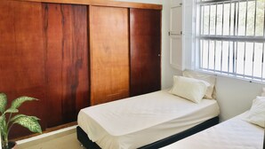 Une chambre avec une armoire en bois, un lit avec une literie blanche, une plante en pot et une fenêtre avec des barreaux.