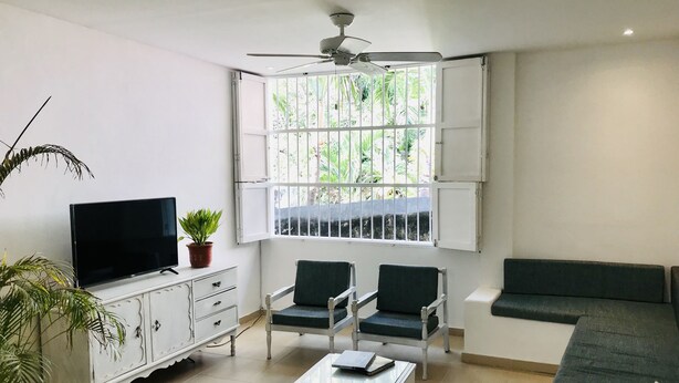Un salon comprenant un téléviseur, deux fauteuils, un canapé, une table basse et une fenêtre avec des barreaux.