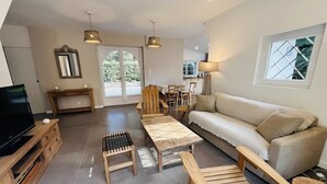 Un salon moderne avec un canapé, une table basse en bois et des fauteuils. On y trouve un téléviseur posé sur un meuble en bois, un coin repas avec des chaises et une grande fenêtre donnant sur un paysage verdoyant.