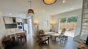 Une cuisine moderne avec des meubles blancs, une table à manger et des chaises. Le salon comprend un canapé et une table basse. Une grande fenêtre offre une vue sur le jardin arrière.