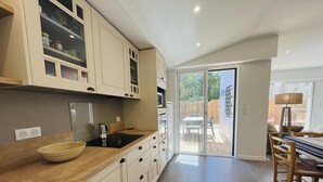 Une cuisine moderne avec un plan de travail en bois, un évier et des placards. On aperçoit un coin repas avec une table et des chaises à travers une porte-fenêtre coulissante qui donne sur une terrasse.