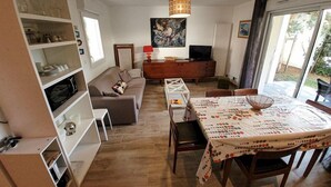 Un salon comprenant un canapé, une table à manger et un téléviseur. On y trouve également des étagères remplies d’ustensiles de cuisine et un tableau accroché au mur.