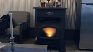 Un salon avec un poêle à bois, une table ornée d’une bougie et d’une décoration en forme de flocon de neige, et un fauteuil.