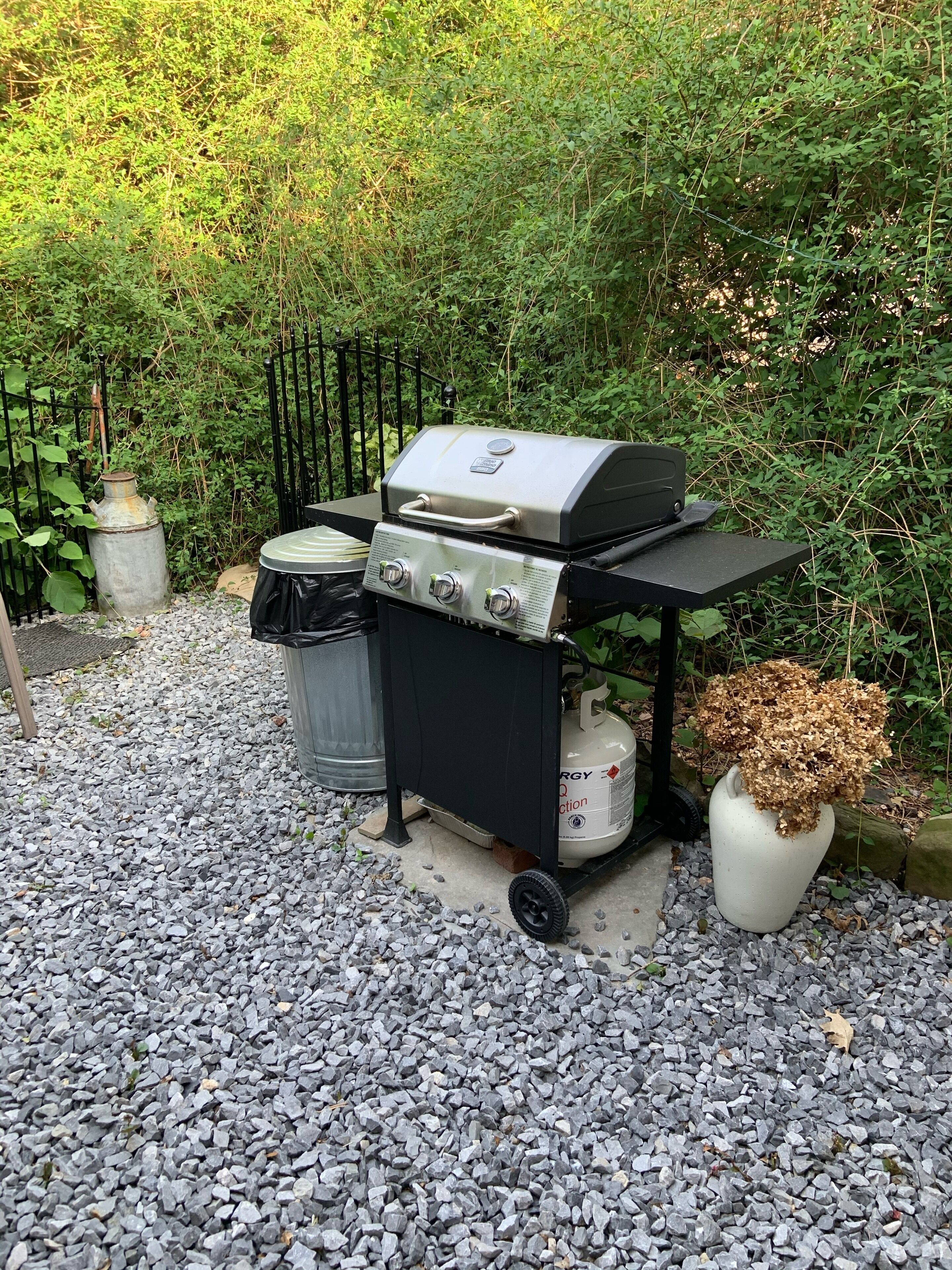 A portable grill with a gas tank and a propane container, set on a gravel surface with a metal fence and greenery in the background.