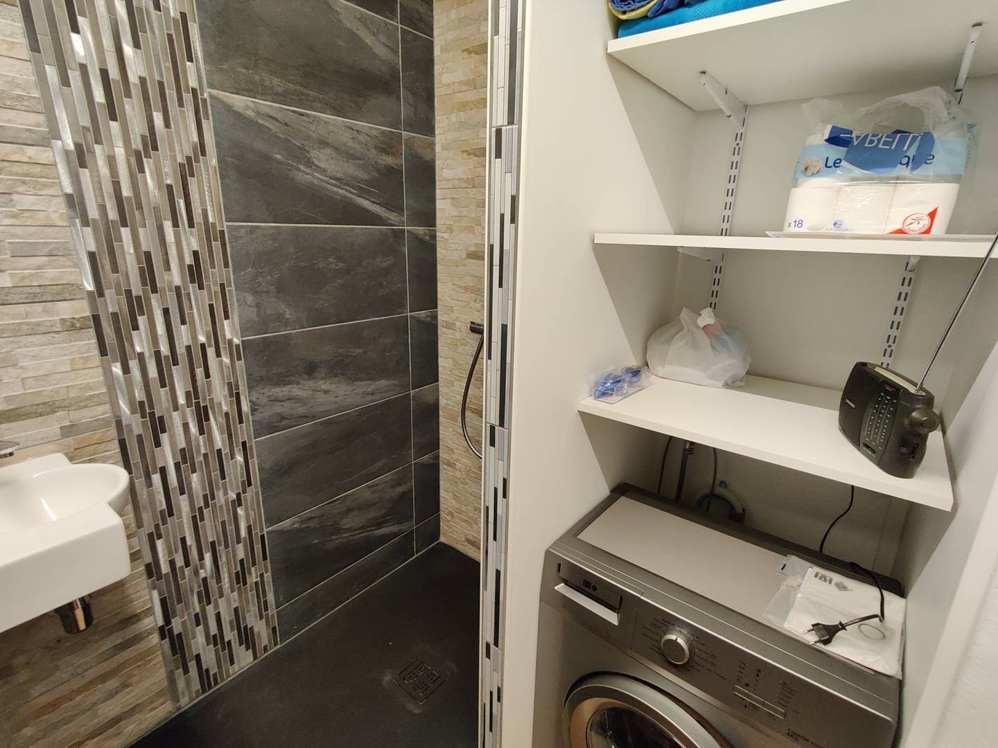 A compact bathroom with a modern design, featuring a shower area with dark tiles and a patterned wall. There is a washing machine and a shelf with toiletries.
