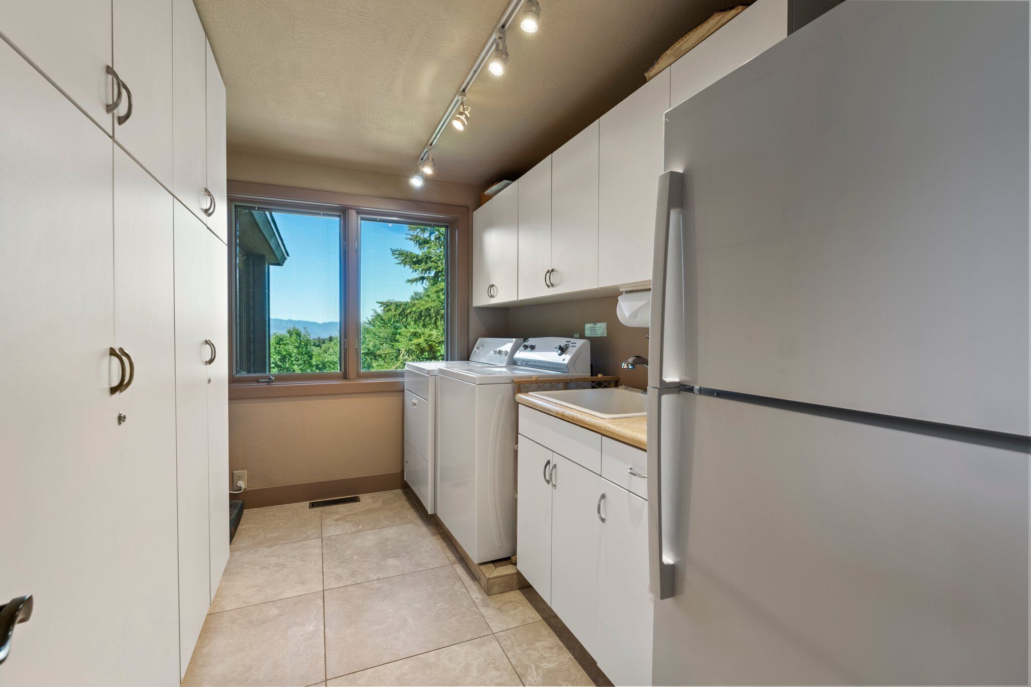 Cocina compacta con armarios blancos, refrigerador de acero inoxidable, lavadora y ventana con vista a la vegetación.