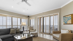 A spacious living room with a grey sofa, a wicker armchair, a glass coffee table, and a large window with blinds. - SeaChase 1601W by Liquid Life (Orange Beach)